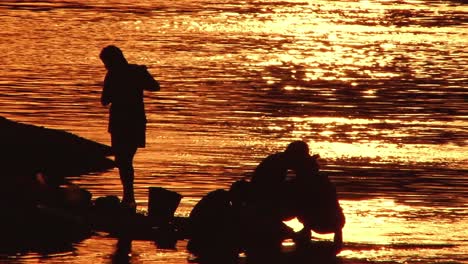 Teleaufnahme-Einer-Familie,-Die-In-Einem-Fluss-Badet-Und-Kleidung-Wäscht,-Der-Durch-Den-Sonnenuntergang-Silhouettiert-Wird