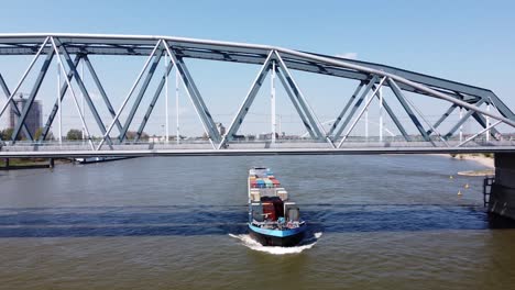 Containerschiff-Fährt-Unter-Brücke-Am-Fluss-Waal-In-Nijmegen,-Gelderland,-Niederlande---Luftschwenk-Links