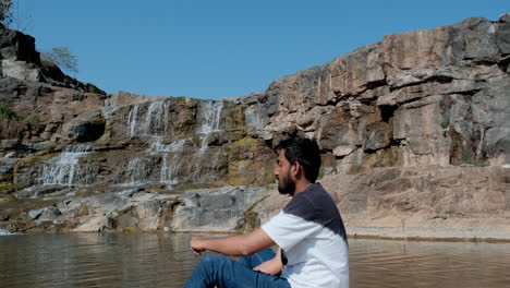 Bärtiger-Mann,-Der-In-Der-Nähe-Eines-Wasserfalls-Sitzt-Und-Steine-In-Den-Fluss-Wirft