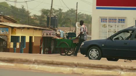 Toma-En-Cámara-Lenta-De-Un-Vendedor-Callejero-De-Café-Empujando-Su-Carro-Con-Autos-Y-Motocicletas-Pasando-En-Primer-Plano---Lomé,-Togo,-áfrica