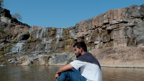 Hombre-Barbudo-Cansado-Sentado-Junto-Al-Río-Cerca-De-La-Cascada-En-Un-Día-Soleado