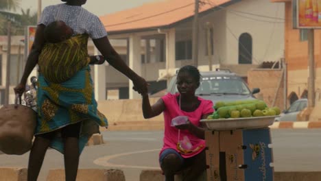 Toma-En-Cámara-Lenta-De-4k-De-Una-Vendedora-Ambulante-De-Agua-Africana-Que-Lleva-A-Un-Bebé-En-La-Espalda-Y-Comercia-Con-Un-Vendedor-De-Frutas,-Togo,-áfrica