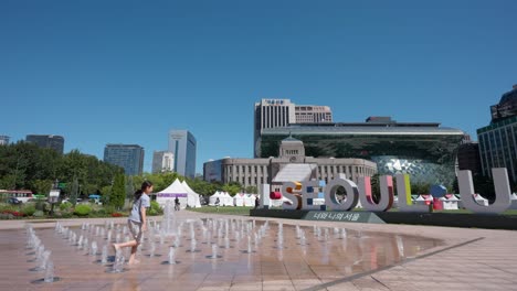 Joven-Coreana-Disfruta-Del-Juego-De-Agua-De-La-Fuente-En-El-Ayuntamiento-De-Seúl-Junto-A-La-Escultura-Simbólica-I-Seoul-U