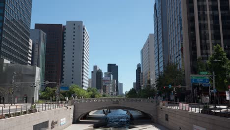 Parque-Del-Arroyo-Cheonggyecheon-Con-Puente-Y-Vista-De-Los-Exteriores-De-Los-Edificios-De-Oficinas-De-Vidrio-Alto-Contra-El-Cielo-Azul---Espacio-De-Copia