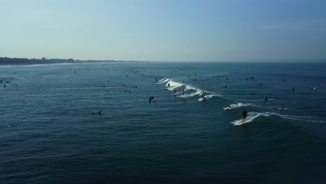 Drone-footage-shows-a-large-number-of-surfers-riding-a-wave-or-swell-while-numerous-individuals-are-swimming-and-floating-in-the-water-near-Pantai-Batu-Bolong,-Canggu-beach