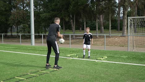 Young-soccer-player-being-trained-by-trainer,-Kicking-the-ball-back-and-tapping-the-floor-a-few-times