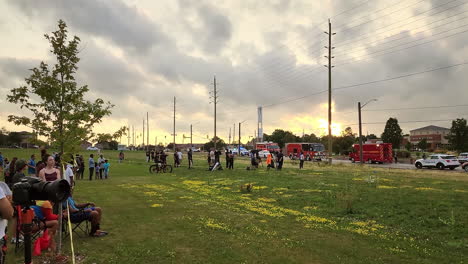 People-watching-from-grass-field-a-crime-scene-with-Firefighters-vehicles-parked-along-the-road,-Panning-shot