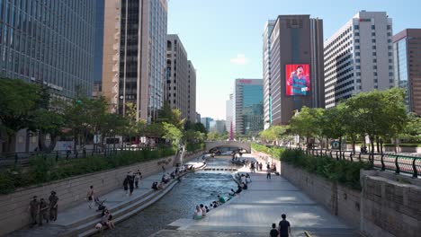 Parque-Del-Arroyo-Cheonggyecheon-Rodeado-De-Rascacielos-De-Oficinas-De-Gran-Altura-Con-Gente-Haciendo-Turismo-En-El-Centro-De-Seúl-O-Descansando-Junto-Al-Agua-Del-Río