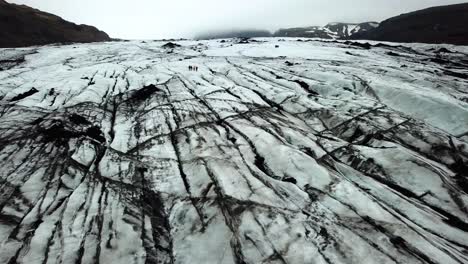 Vista-Aérea-Del-Paisaje-De-Personas-Caminando-En-El-Hielo-Texturizado-Del-Glaciar-Sólheimajökull,-Islandia,-En-Verano