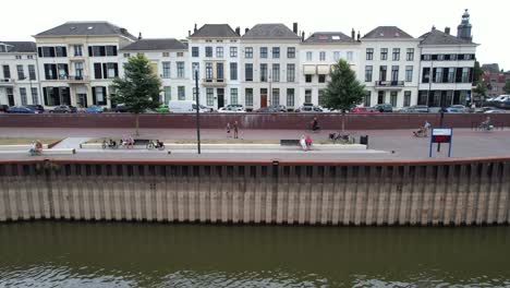 Seitliche-Luftbewegung,-Die-Menschen-Zeigt,-Die-Freizeitaktivitäten-Auf-Dem-Ijsselkade-Boulevard-Der-Turmstadt-Zutphen-Ausüben,-Mit-Niedrigem-Wasserstand-Des-Flusses-Ijssel-Im-Vordergrund