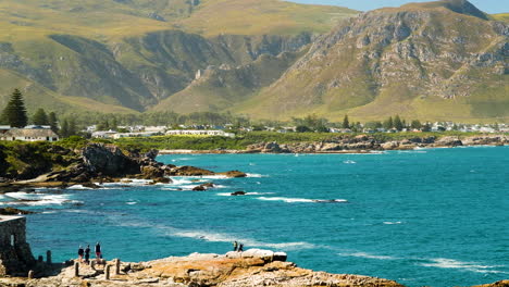 Malerische-Küste-Von-Hermanus-Mit-Wunderschönen-Bergen-Im-Hintergrund---Touristen,-Die-Von-Den-Felsen-Aus-Wale-Beobachten