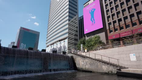 Cascada-Del-Arroyo-Cheonggyecheon-Y-Gran-Cartelera-Digital-De-Anuncios-Públicos-Reproduciendo-Comerciales-De-Airpods-En-Un-Hermoso-Día-De-Verano-Mientras-La-Gente-Camina-En-La-Plaza-Cheonggye-En-La-Ciudad-De-Seúl