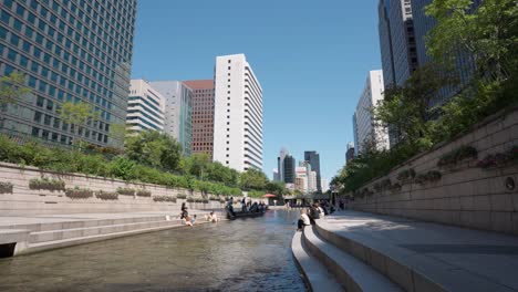 Gente-Descansando-Sentada-Junto-Al-Arroyo-Cheonggyecheon-En-Un-Caluroso-Día-De-Verano-En-El-Centro-De-La-Ciudad-De-Seúl-Con-Vistas-A-Los-Altos-Rascacielos,-Corea-Del-Sur