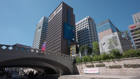 La-Gente-Se-Esconde-Bajo-El-Puente-A-La-Sombra-En-Un-Caluroso-Día-De-Verano-Descansando-Junto-Al-Arroyo-Cheonggyecheon-En-El-Centro-De-La-Ciudad-De-Seúl-Con-Vistas-A-Los-Rascacielos-De-La-Torre-Alta-Contra-El-Cielo-Azul