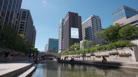 Waterfront-view-of-Cheonggyecheon-Stream-Park-with-people-tourists-resting-by-the-water-on-Summer-Beautiful-Day-in-Seoul-urban-landscape,-South-Korea