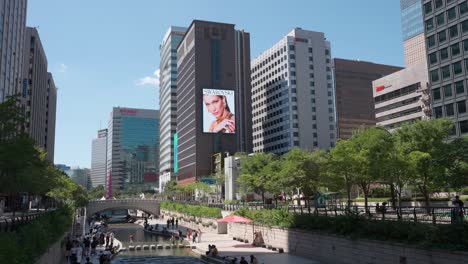 La-Gente-Viaja-En-El-Parque-Del-Arroyo-Cheonggyecheon-En-La-Ciudad-De-Seúl,-Swarowski-Y-El-Anuncio-Comercial-De-Audi-En-Una-Enorme-Pantalla-Digital-De-Pared-En-El-Centro