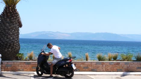 Un-Hombre-Con-Gafas-De-Sol,-Un-Pantalones-Blanco-Y-Un-Pantalón-Corto-Llega-En-Una-Moto-Azul-Y-Se-Estaciona-Frente-A-Una-Palmera-Con-Un-Paisaje-De-Un-Mar-Azul-Profundo-Y-Una-Montaña-Detrás-De-él-En-Un-Día-Soleado-En-Grecia