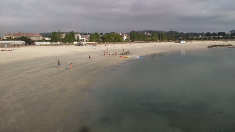 Elderly-woman-strolls-along-the-shore-of-the-beach-with-two-children-playing-soccer-and-a-group-of-people-prepare-to-paddle-on-a-cloudy-morning,-blocked-panoramic-shooting,-Porto-Novo,-Galicia,-Spain