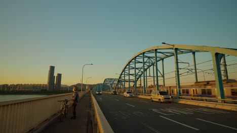 El-Tráfico-Del-Puente-Dongjak-Y-El-Tren-De-La-Línea-4-Del-Metro-Pasan-Al-Atardecer,-Un-Ciclista-Irreconocible-Con-Una-Bicicleta-Parada-Para-Disfrutar-De-La-Vista-De-La-Puesta-De-Sol-Desde-La-Pasarela-Del-Puente,-Seúl,-Corea-Del-Sur