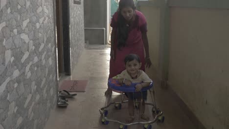 Pretty-Asian-baby-boy-learning-to-baby-walk-and-strolling-front-in-baby-walker-outside-in-the-corridor-beside-his-mother