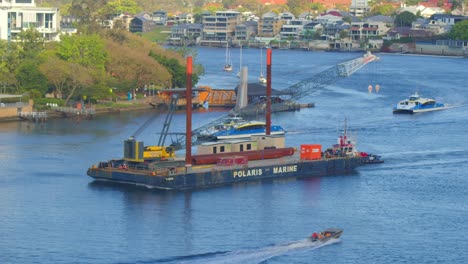 Ein-Großer-Lastkahn-Mit-Einem-Kran-Segelt-Den-Fluss-Hinauf,-Flankiert-Von-Stadtkatamaranen