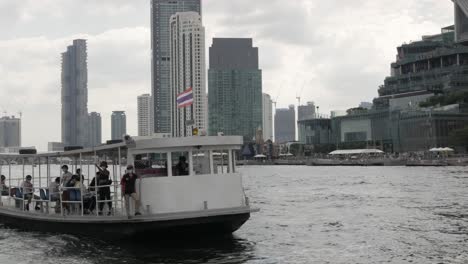 Vista-Panorámica-Del-Ferry-De-Pasajeros-En-El-Río-Chaophraya-Con-Un-Edificio-De-Hotel-Rascacielos-En-La-Orilla-Del-Río