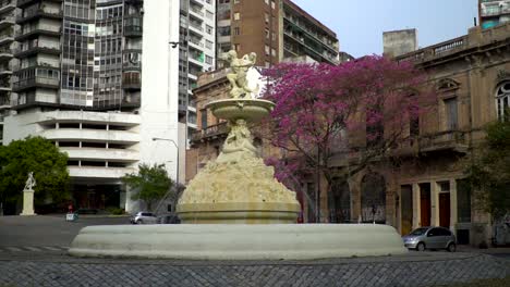 Cámara-Lenta,-Vista-Panorámica-Manual-De-La-Fuente-De-Las-Utopías-En-Rosario,-Argentina,-Contra-Un-Hermoso-Telón-De-Fondo-De-Edificios-Y-Un-árbol-De-Jacaranda-Florecido