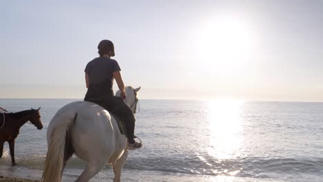 Rückansicht-Des-Reiters-Mit-Helm,-Der-Bei-Sonnenuntergang-Auf-Pferden-Ins-Meerwasser-Reitet---Genießen-Sie-Im-Sommer-Ein-Gutes-Gefühl-Unter-Wasser