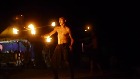 Asian-male-spinning-dual-handed-fire-poles-in-large-circular-motion-while-performing-acrobatic-tricks,-filmed-in-handheld-style