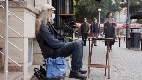 An-elderly-man-in-a-face-mask-sitting-on-the-edge-of-a-sidewalk-with-his-back-to-the-street