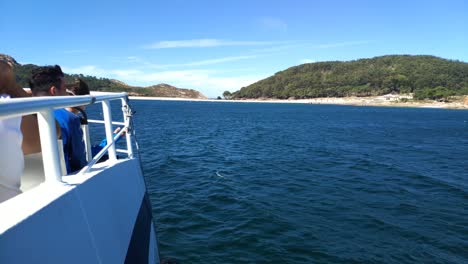 Lado-Del-Barco-Con-Turistas-Navegando-Frente-A-Las-Islas-Cíes-Con-Sus-Colinas,-Playa-Y-Bosque-Un-Día-Soleado-De-Cielo-Azul,-Tiro-Bloqueado,-Pontevedra,-Galicia,-España