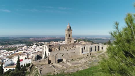 Luftwagen-Schoss-Durch-Bäume-Zur-Historischen-Kirche-Von-Santa-Maria-In-Medina-Sidonia-In-Spanien-In-Der-Provinz-Cadiz-Mit-Blick-Auf-Die-Altstadt-Mit-Weißen-Häusern-An-Einem-Sonnigen-Tag