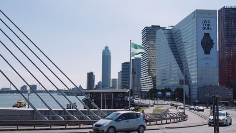 Toma-En-Cámara-Lenta-De-Autos-Conduciendo-A-Lo-Largo-Del-Puente-Con-El-Edificio-Witte-Huis-En-El-Fondo,-Bandera-De-Rotterdam-Ondeando