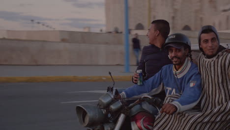 Tres-Hombres-árabes-Felices-En-Una-Motocicleta-De-Tres-Ruedas-De-Entrega-Conducen-En-La-Rotonda-Frente-A-La-Mezquita-Hassan-ii-En-Casablanca-Marruecos-Y-Cabalgan-Hacia-La-Puesta-De-Sol-A-Lo-Largo-De-La-Costa-Atlántica