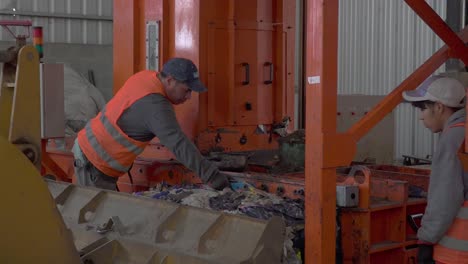 A-pair-of-workers-load-compacted-garbage-onto-a-bulldozer-inside-a-waste-processing-plant