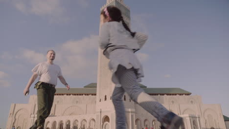 Military-aged-man-walking-confidently-crosses-path-with-happy-little-girl-running-in-front-of-Hassan-II-mosque-in-Casablanca-Morocco