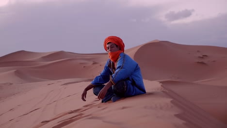 Orgulloso-Hombre-Marroquí-Bereber-Mirando-A-La-Cámara-Y-En-La-Distancia,-Sentado-En-Las-Dunas-De-Arena-En-Erg-Chubby,-Merzouga,-Marruecos