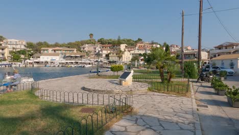 Vista-Del-Monumento-Y-Del-Paseo-Marítimo-De-Kassiopi,-Gente-Sentada-En-El-Banco-A-Orillas-Del-Mar