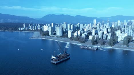 barge-afloat-on-beach-aerial-180-degree-view-of-boats-passing-by-as-workers-dismantle-cargo-ship-and-crane-sorting-metals-into-container-for-repurpose-use-a-year-later-on-a-sunny-day-in-English-Bay