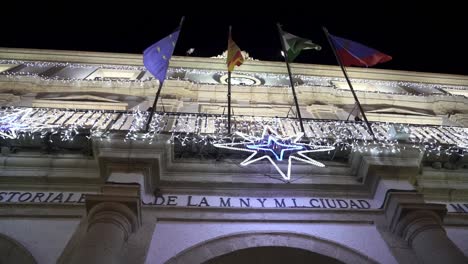 Tiro-De-Cámara-Lenta-De-Paralaje-De-ángulo-Bajo-Del-Ayuntamiento-Casa-Consistorial-En-Medina-Sidonia-Provincia-De-Cádiz-En-España-Con-Balcón-Decorado-Con-Luces-De-Hadas-Y-Elementos-Navideños-Y-Diferentes-Banderas