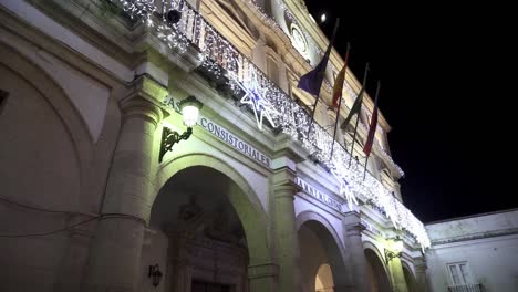 Tiro-Inclinado-En-Cámara-Lenta-Del-Ayuntamiento-Casa-Consistorial-En-Medina-Sidonia-En-Cádiz-España-Con-Balcón-Decorado-Para-Navidad-Con-Luces-De-Hadas-Y-Elementos-Navideños-Y-Diferentes-Banderas