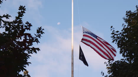 American-Flag-At-Half-Mast-Waving-Beside-Official-Emblem-of-National-League-of-Families-POW-MIA-Flag-in-Central-Park,-Low-Angle-View-Through-Trees,-Memorial-Day