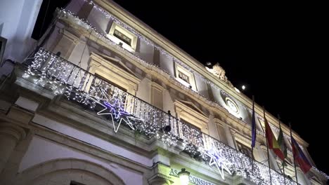 Toma-En-Cámara-Lenta-De-ángulo-Bajo-Del-Ayuntamiento-Casa-Consistorial-En-Medina-Sidonia-Cádiz-Decorada-Con-Elementos-Navideños-Y-Luces-De-Hadas-Y-Banderas-De-Diferentes-Países