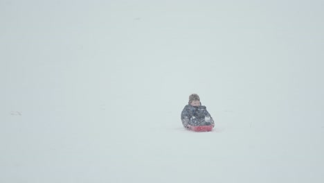 Niño-Deslizándose-Por-La-Ladera-De-Una-Montaña-Con-Mucha-Nieve-Cayendo-Activamente