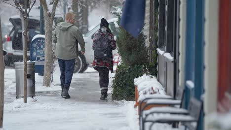 Mann-Und-Frau-Gehen-Den-Bürgersteig-Hinunter-Und-Zeigen-Auf-Die-Schaukästen-Von-Schaufenstern-Mit-Schnee-Draußen