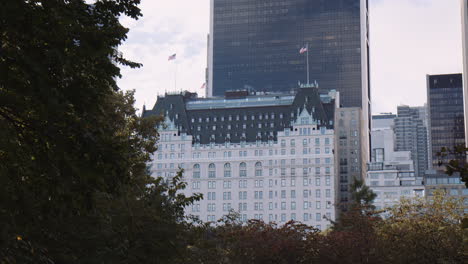 Vista-Del-Edificio-Del-Hotel-Plaza-Desde-El-Otro-Lado-Del-Parque-Central,-La-Ciudad-De-Nueva-York,-Manhattan,-Ee.uu.,-La-Vegetación-De-Los-árboles-Del-Parque-Urbano-En-Primer-Plano-Y-Las-Torres-Modernas-De-Los-Edificios-Del-Centro-En-El-Fondo