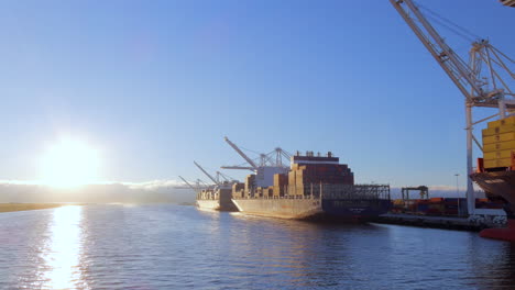 Vista-Aérea-De-ángulo-Bajo-Que-Pasa-Por-Portacontenedores-Del-Puerto-De-Oakland-Y-Grúa-De-Pórtico-Industrial-En-El-Reluciente-Paseo-Marítimo