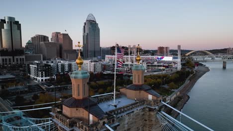 Luftaufnahme-Der-Flagge-Der-Roebling-Brücke-Mit-Dem-Stadtbild-Von-Cincinnati-Im-Hintergrund---Zeitlupe,-Umlaufbahn,-Drohnenaufnahme