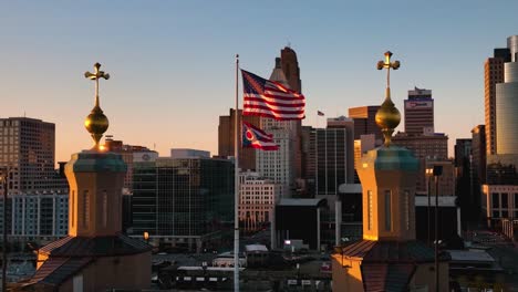 Vista-Aérea-Alrededor-De-La-Bandera-De-Ohio-Y-Estados-Unidos-Ondeando-Con-El-Fondo-De-La-Ciudad,-Puesta-De-Sol-En-Estados-Unidos---Cámara-Lenta