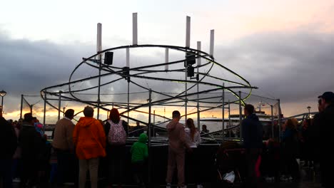 Public-interact-with-illuminated-spiral-light-looper-neuron-artwork,-Liverpool-pier-head-river-of-light-event-with-dramatic-sunset